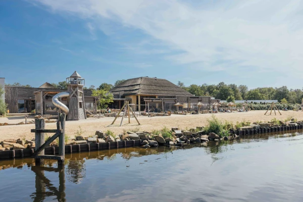 Uitzicht vanaf het water op het speelstrand en centrumgebouw Ganvie Village van Lake Resort Beekse Bergen
