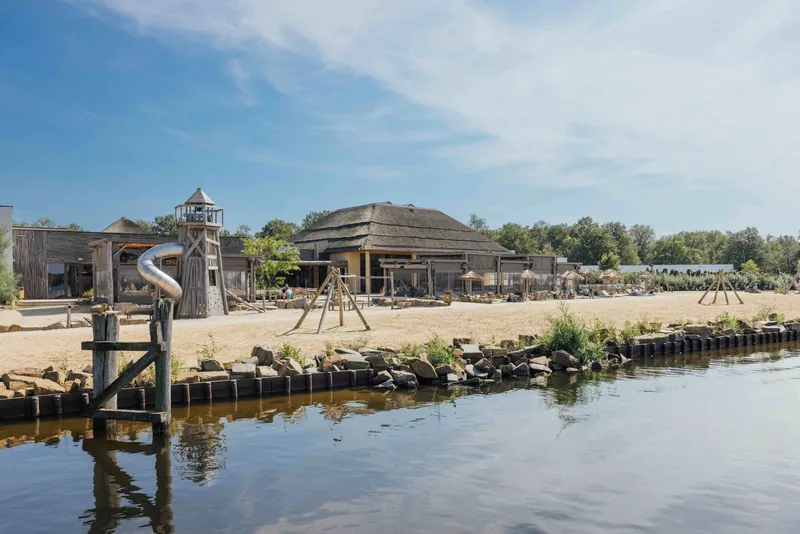 Uitzicht vanaf het water op het speelstrand en centrumgebouw Ganvie Village van Lake Resort Beekse Bergen