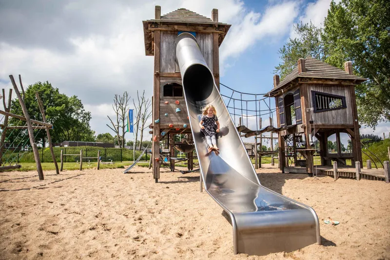Een meisje glijdt van de hoge metalen glijbaan van het houten speelkasteel in de zandspeeltuin van het recreatiepark.
