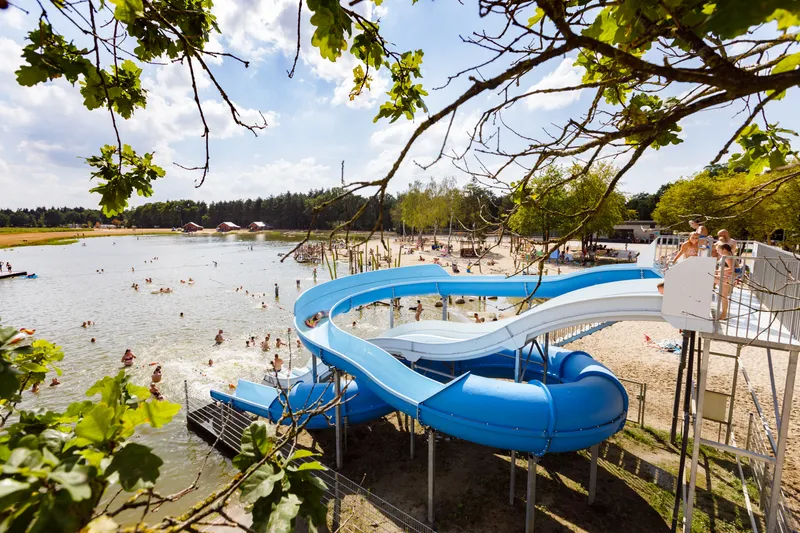 Kinderen glijden van de grote blauwe waterglijbaan het water in bij recreatieplas 't Witven op het zandstrand van Molecaten Park De Flaasbloem.