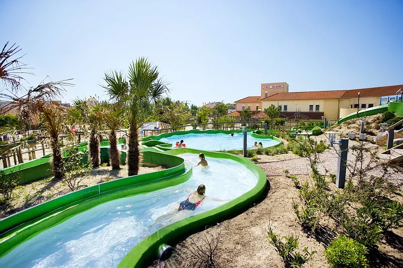 Kinderen glijden door het buitengedeelte van de wildwaterbaan van de Aqua Mundo op Center Parcs Port Zélande, omringd door palmbomen en duinlandschap.