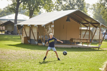 Een jongen die voor zijn bruine safaritent aan het voetballen is op het gras.