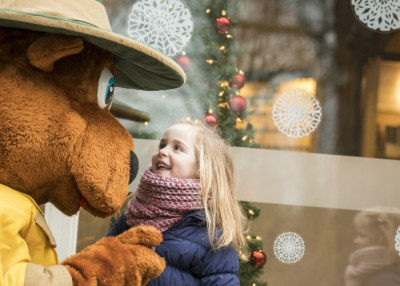 Een lachend meisje kijkt op naar een vriendelijke mascottefiguur Bollo en met een kerstversierde achtergrond in Landal