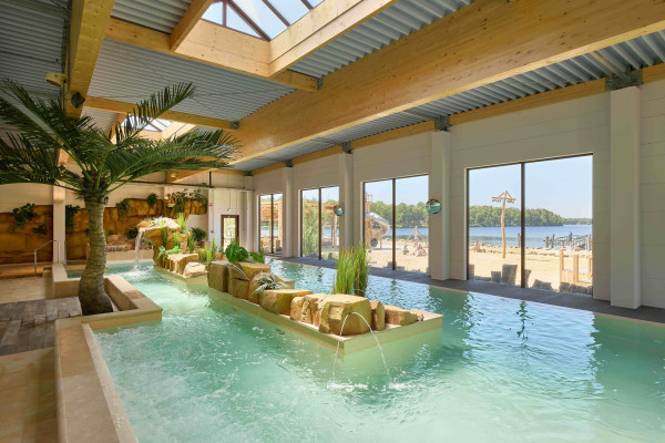 Binnenzwembad in tropische sferen met palmbomen en uitzicht door grote ramen op het zandstrand en het meer