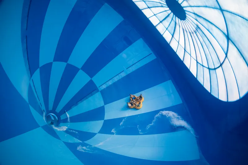 Een groep mensen in een gele opblaasbare band glijdt door de gigantische, blauw-wit geblokte trechter van de waterglijbaan Saltato in het zwemparadijs van Landal Hof van Saksen.