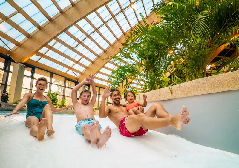 Lachende familie met jonge kinderen glijdt samen van de brede familieglijbaan in het overdekte zwemparadijs van camping Domaine des Ormes.