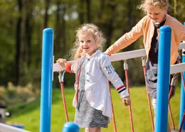 Een meisje met blonde krullen balanceert lachend over een wiebelige touwbrug in de buitenspeeltuin van Recreatiepark Beekbergen.
