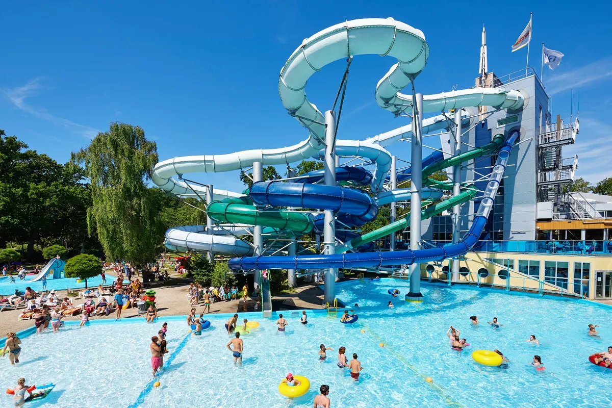 Het buitengedeelte van het Tikibad op Duinrell, met een groot complex van witte en groene waterglijbanen en een vol zwembad op een zonnige dag.