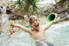 Een lachende jongen met zijn armen wijd heeft waterpret in het zwembad van de Aqua Mundo bij Center Parcs Erperheide, met andere kinderen op de achtergrond.
