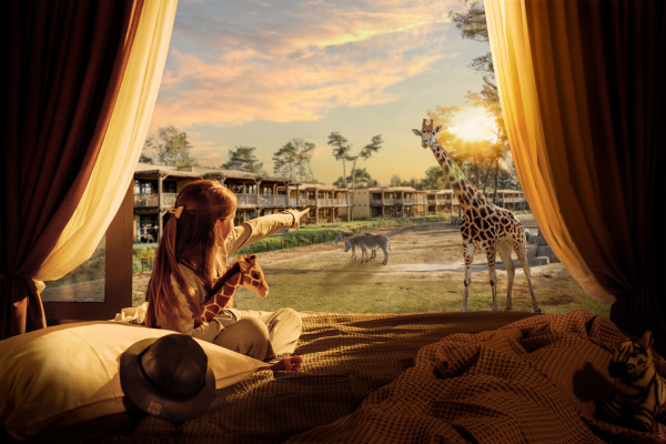 Een meisje met een knuffelgiraffe in haar arm wijst vanuit haar lodge enthousiast naar de echte giraffen buiten bij safari hotel beekse bergen