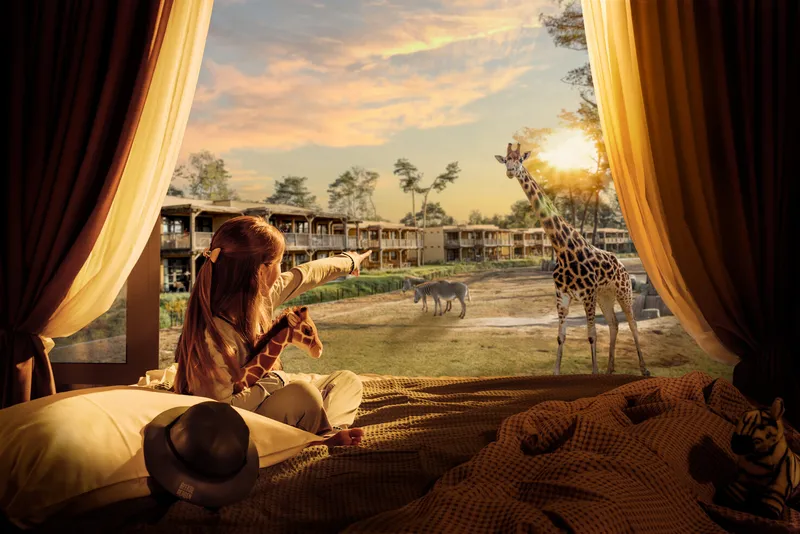  Een meisje met een knuffelgiraffe in haar arm wijst vanuit haar lodge enthousiast naar de echte giraffen buiten bij safari hotel beekse bergen