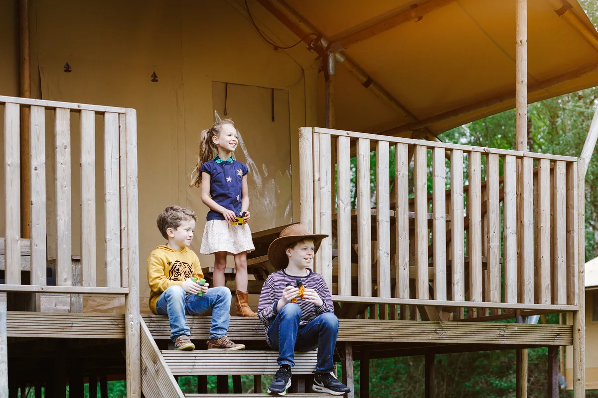 Drie kinderen zitten op de houten veranda van een safaritent op Vakantiepark Beekse Bergen, klaar voor avontuur.