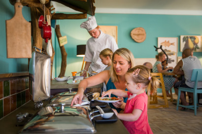 Een moeder helpt haar jonge dochter met het opscheppen van eten bij een kinderbuffet in het restaurant van Landal Sluftervallei, met een chef-kok op de achtergrond.