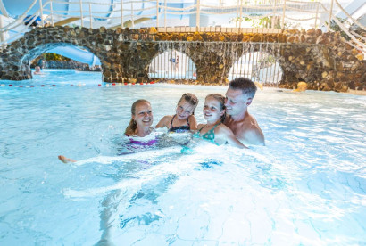 Een lachend gezin met vader en drie dochters heeft plezier in het water onder een brug met waterval in het subtropische zwemparadijs van Marveld Recreatie.