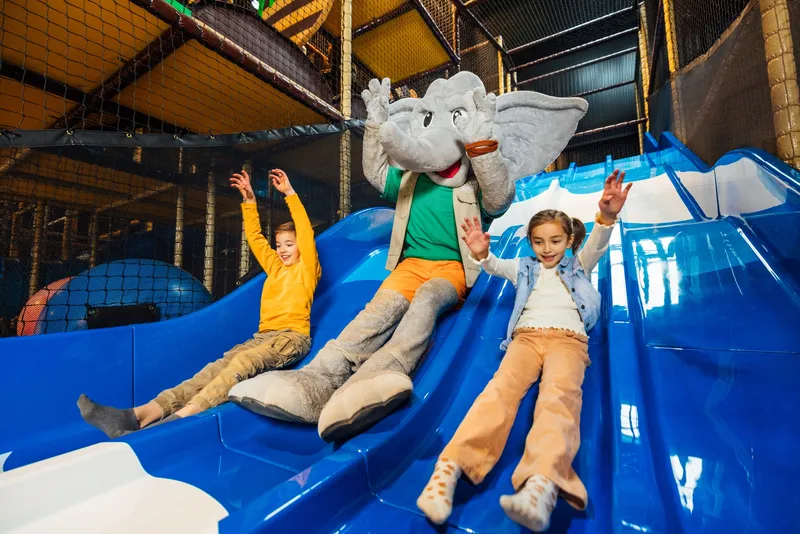 Twee kinderen glijden lachend van een blauwe glijbaan met mascotte Djambo in Speelland Beekse Bergen