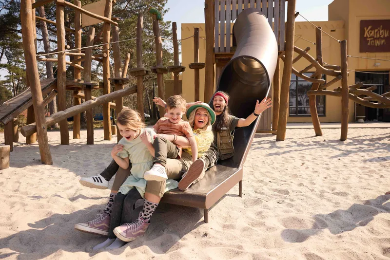 Vrolijk gezin met twee jonge kinderen glijdt samen lachend van de glijbaan in de buitenspeeltuin op vakantiepark Beekse Bergen