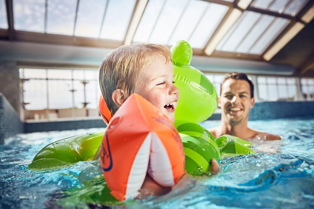 Een lachende peuter met zwemvleugels speelt samen met zijn vader in het binnenzwembad van Vakantiepark De Krim.