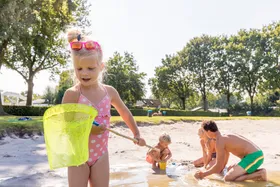 Een meisje in een roze badpak staat met een schepnetje aan de rand van de zanderige speelvijver op Recreatiepark De Leistert.