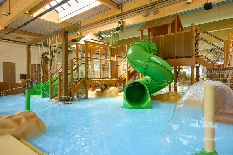 Overdekt zwemparadijs met een grote groene waterglijbaan en diverse houten speeltoestellen
