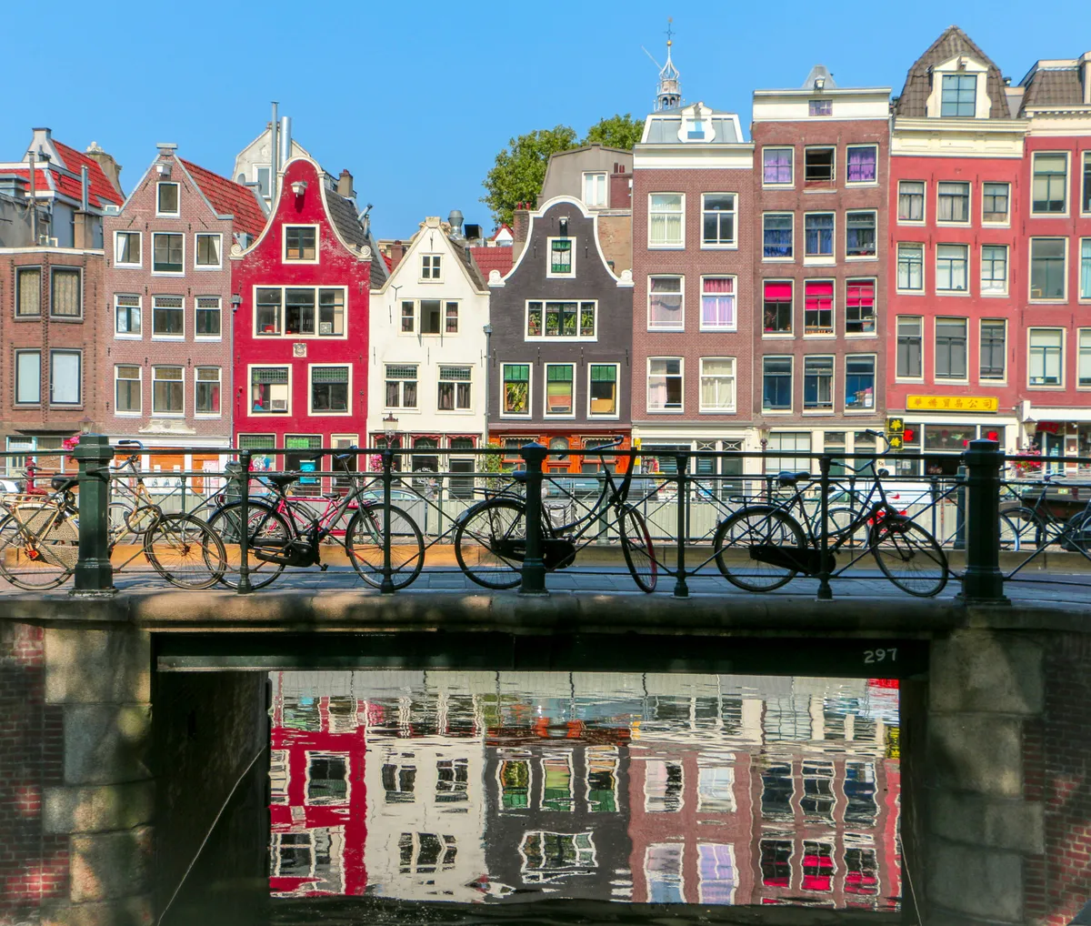 Gekleurde grachtenpanden in Amsterdam weerspiegelend in het water, met fietsen geparkeerd op de brug op de voorgrond.