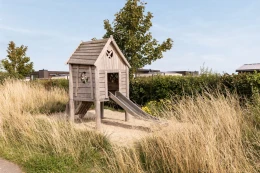 Houten speeltoestel met een glijbaan in een natuurspeeltuin met hoog gras op Vakantiepark Zeedijk in Zuid-Holland.
