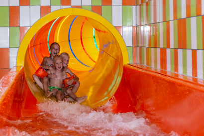 Drie kinderen komen met een grote plons uit de oranje tunnelglijbaan in het binnenzwembad van Recreatiecentrum De Vogel.
