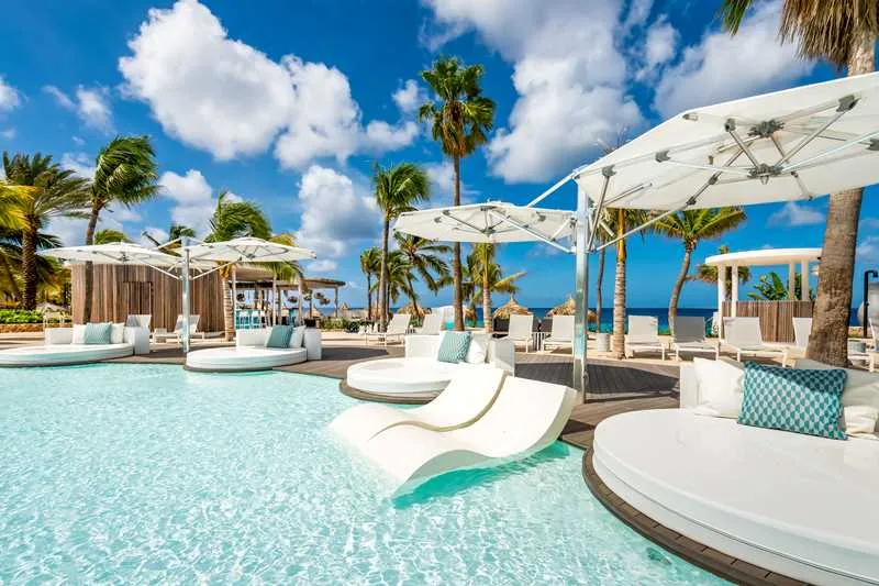 Luxe resort met een zwembad vol witte design ligbedden en uitzicht op een tropisch strand met palmbomen.