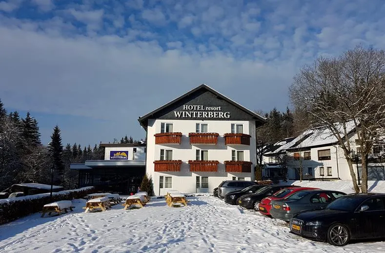 De buitenkant van Hotel Winterberg Resort op een zonnige winterdag, met een besneeuwde parkeerplaats en terras.
