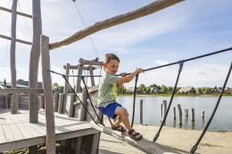 Een jongen balanceert behendig over een houten touwbrug die deel uitmaakt van het klimparcours boven de recreatieplas van Hof van Saksen.