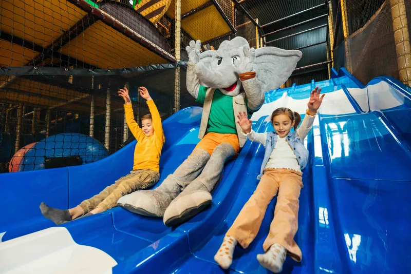Twee kinderen glijden lachend van een blauwe glijbaan met mascotte Djambo in Speelland Beekse Bergen