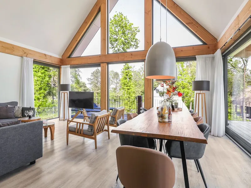 Ruime en lichte woonkamer van een luxe vakantievilla op Landal Puur Exloo, met een hoog plafond, houten balken, en enorme ramen die een prachtig uitzicht bieden op de omringende natuur en het bos.
