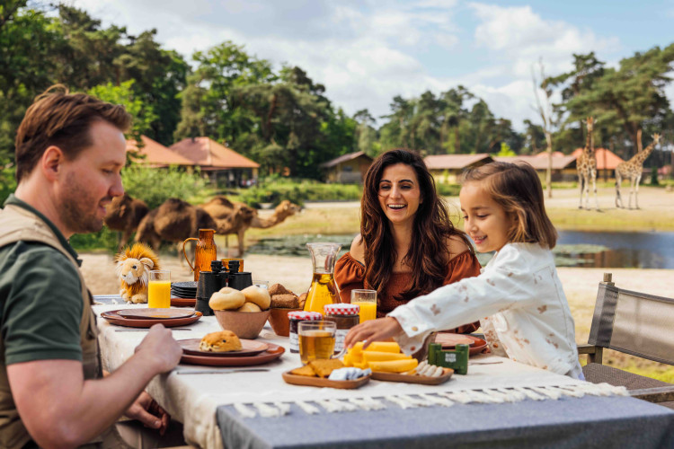 Gezin met kind ontbijt op het terras van hun vakantiehuisje met uitzicht op kamelen en giraffen op Safari Resort Beekse Bergen