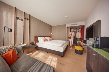 Vrouw loopt binnen in moderne hotelkamer in ATLANTIC Hotel Münster met tweepersoonsbed en een zithoek