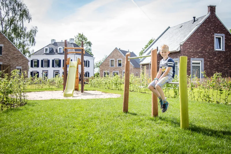 Een jongen speelt op een speeltoestel in een groene, parkachtige speeltuin tussen de vakantiehuizen van Dormio Resort Maastricht.
