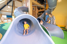 Jong kind komt lachend uit de tunnelglijbaan in de binnenspeeltuin, terwijl ouders toekijken bij Landal Orveltermarke.