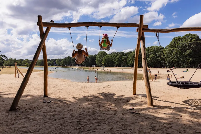 Twee kinderen schommelen naast elkaar op de houten schommels op het zandstrand van Bospark 't Wolfsven in Noord-Brabant.