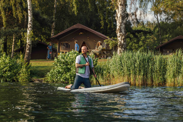 Man met een zwemvest is aan het suppen op het meer met op de achtergrond een safaritent en bomen in Lake Rsort Beekse Bergen