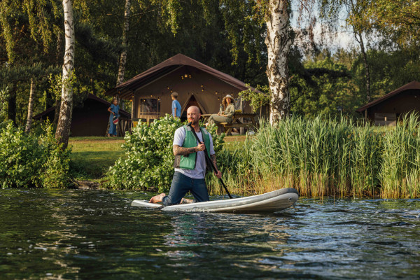 Man met een zwemvest is aan het suppen op het meer met op de achtergrond een safaritent en bomen in Lake Rsort Beekse Bergen