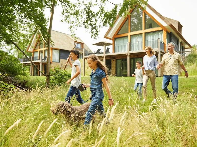 Een familie met kinderen en een hond maakt een wandeling door het hoge gras op vakantiepark Landal Puur Exloo in Drenthe, met op de achtergrond de luxe vakantievilla's.