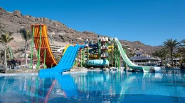 De hoge en kleurrijke waterglijbanen van het Aquapark Lago Taurito, met een brede blauwe en een steile oranje glijbaan die uitkomen in het zwembad.