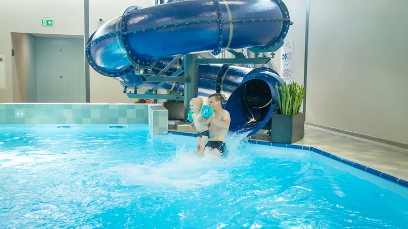 Een vader en zijn jonge kind komen met een grote plons samen van de blauwe waterglijbaan in het binnenzwembad van Dormio Resort Maastricht.