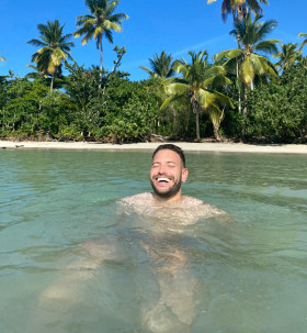 Rolf Sanchez zwemt en lacht in tropische zee met palmbomen op de achtergrond