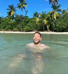 Rolf Sanchez zwemt en lacht in tropische zee met palmbomen op de achtergrond