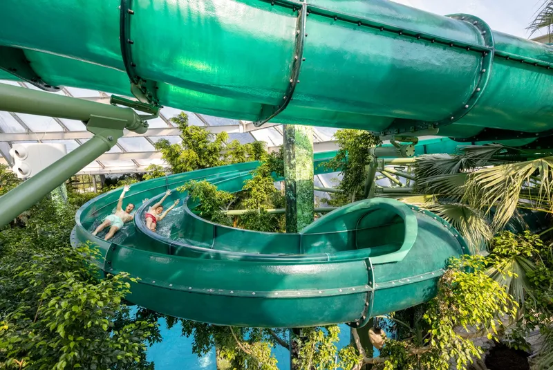 Een bezoeker glijdt met hoge snelheid door een lange, groene en deels doorzichtige waterglijbaan die tussen de tropische planten van Aqua Mundo slingert.