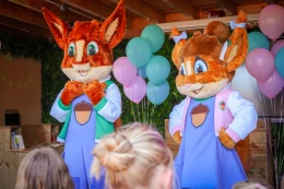 Mascottes Kiki en Koen van De Boshoek staan op een podium voor kids, omringd door kleurrijke ballonnen. 