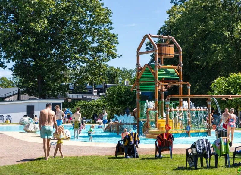 Zonnig overzicht van het buitenzwembad met de grote waterspeeltuin (spraypark) waar kinderen en gezinnen genieten van waterpret op Vakantiepark Westerbergen.
