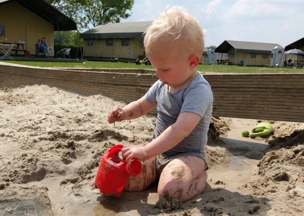 Baby speelt met rode gieter in zandbak met safaritenten op de achtergrond