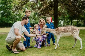 Een compleet gezin met drie kinderen in boeren overalls voert een damhert op het gras bij Vakantiepark Dierenbos.