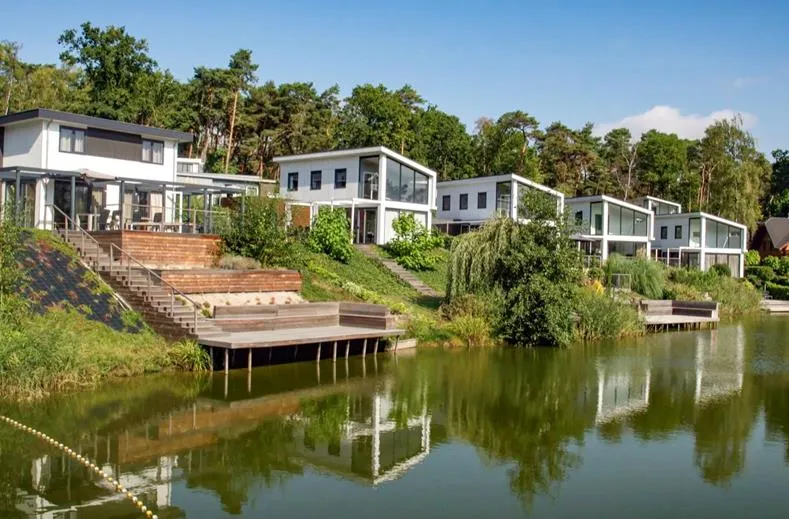Gasten zwemmen en zonnen bij de recreatieplas met zandstrand op Resort Brunssummerheide in Limburg, met uitzicht op de luxe waterwoningen.