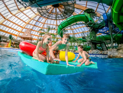 Drie kinderen spelen samen op een grote, drijvende mat in het zwembad van het subtropisch zwemparadijs van Landal De Lommerbergen, met een grote waterglijbaan op de achtergrond.